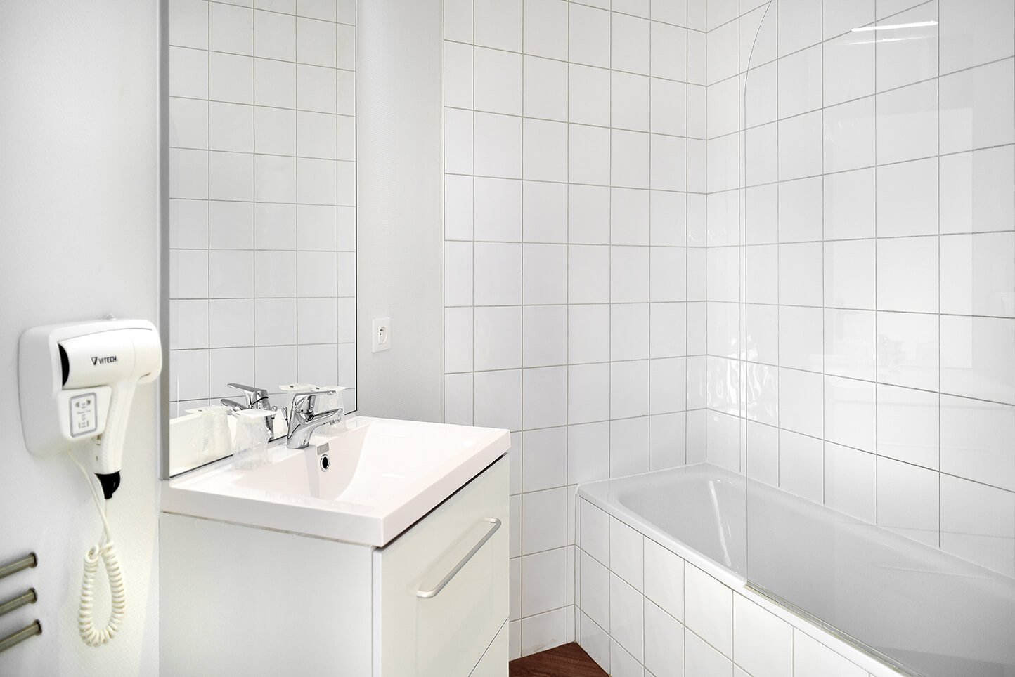 Une salle de bain avec la baignoire sur la droite et le meuble vasque sur la gauche. Au premier plan à gauche, un sèche-cheveux 