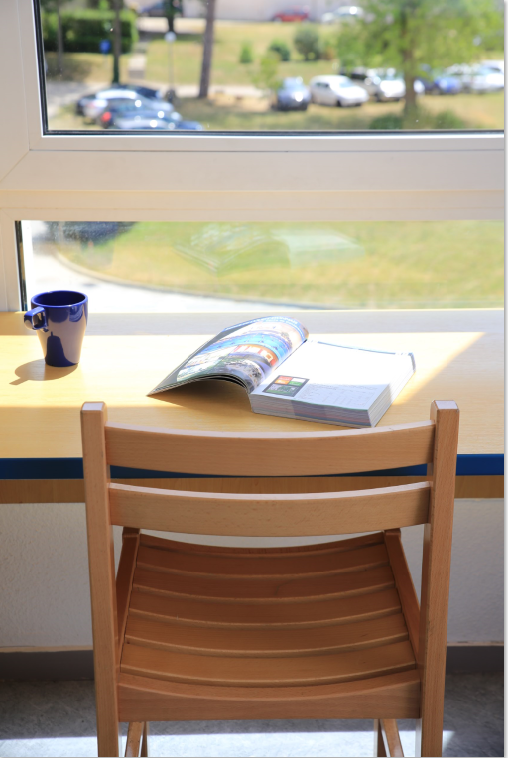 En arrière-plan une fenêtre donnant sur le parc arboré avec devant un bureau ensoleillé sur lequel sont posés une tasse et un livre. Au premier plan une chaise orientée face à la vue 